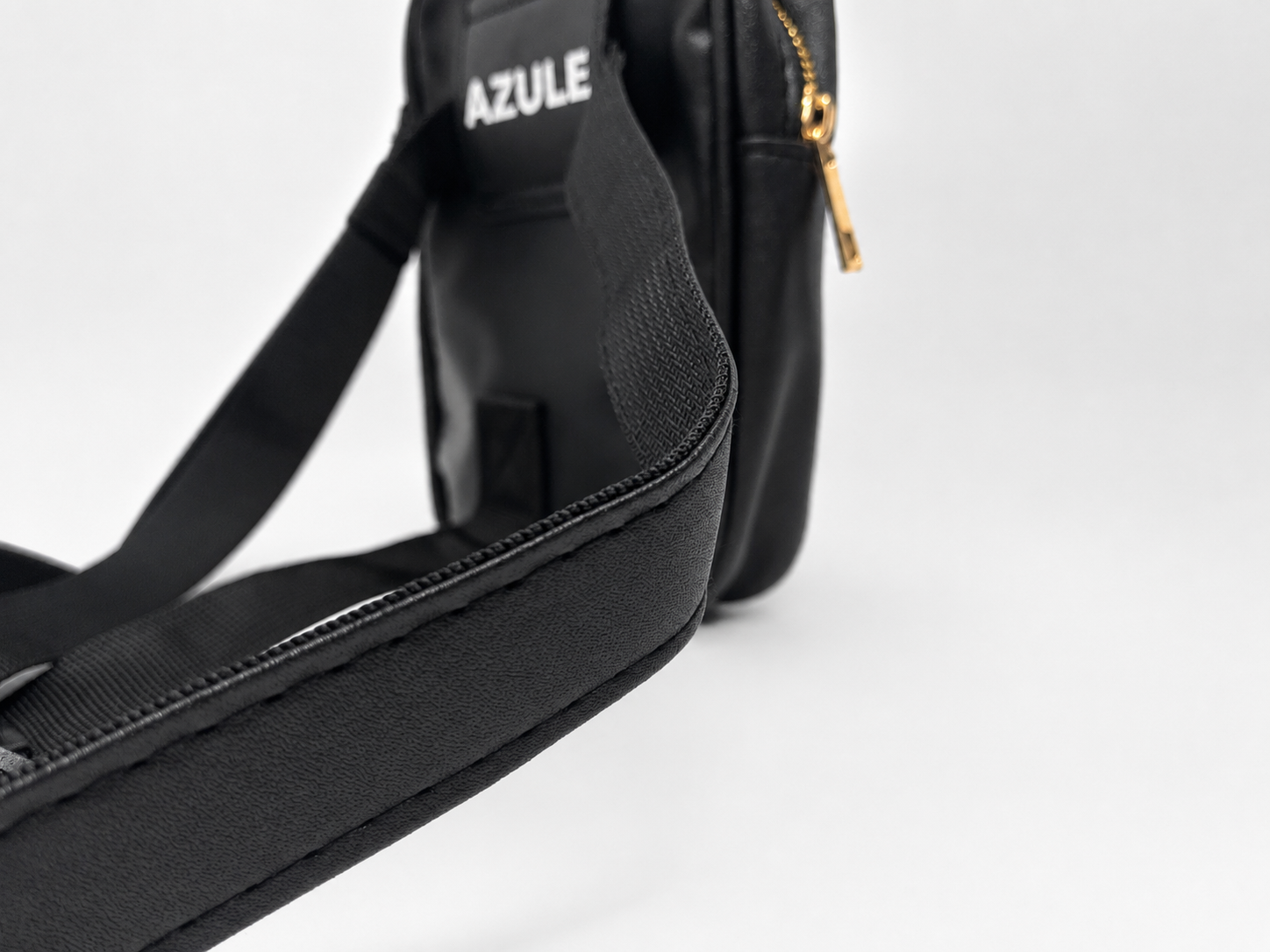 Close-up of a black Azule crossbody bag showing the textured fabric strap, white brand logo, and a gold zipper pull on a light gray background.